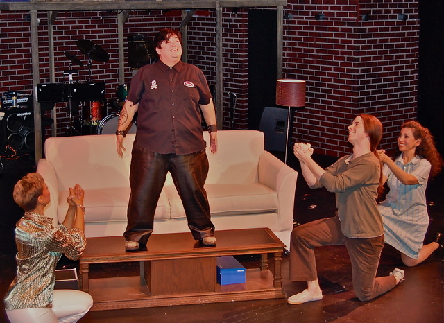 Lea DeLaria standing on a coffee table as the Insatiable Hunger ensemble reacts